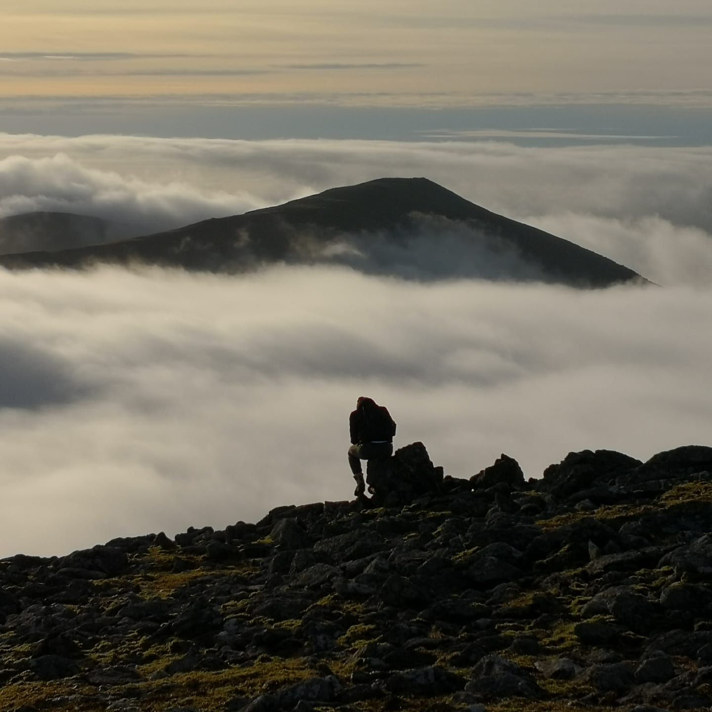 A walker sitting alone on a rocky summit above a sea of cloud, another peak rising through it
